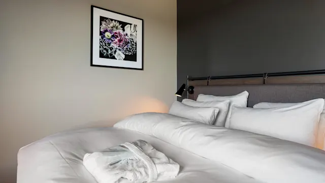 En pent oppredd seng med hvitt sengetøy og en brettet badekåpe hviler under et innrammet blomstermaleri på veggen i et moderne soverom.