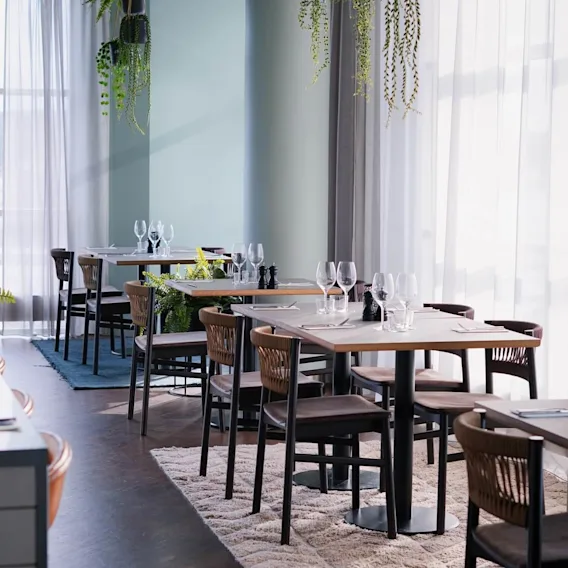Restaurangbord är dukade med glas och bestick, omgivna av växter och badar i naturligt ljus från fönster. Ordet "THE SOCIAL" syns på ett fönster.