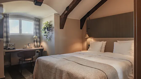 A cozy hotel room with a comfortable bed, a small desk by a window, and rustic wooden beams on the ceiling.