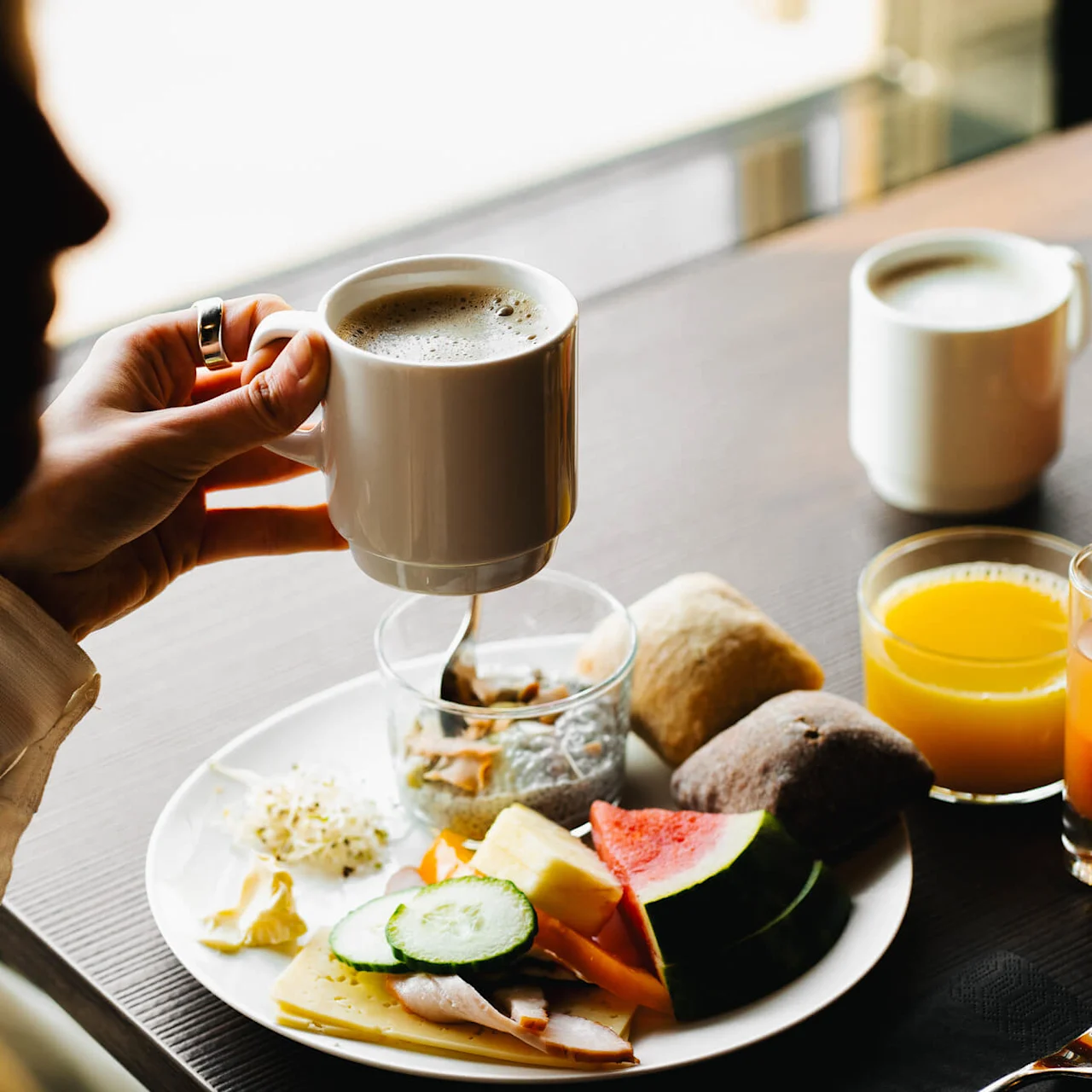 En person holder en kaffekop over en morgenmadstallerken med ost, agurk, vandmelon og brød. Yderligere drikkevarer inkluderer appelsinjuice, placeret på et træbord.