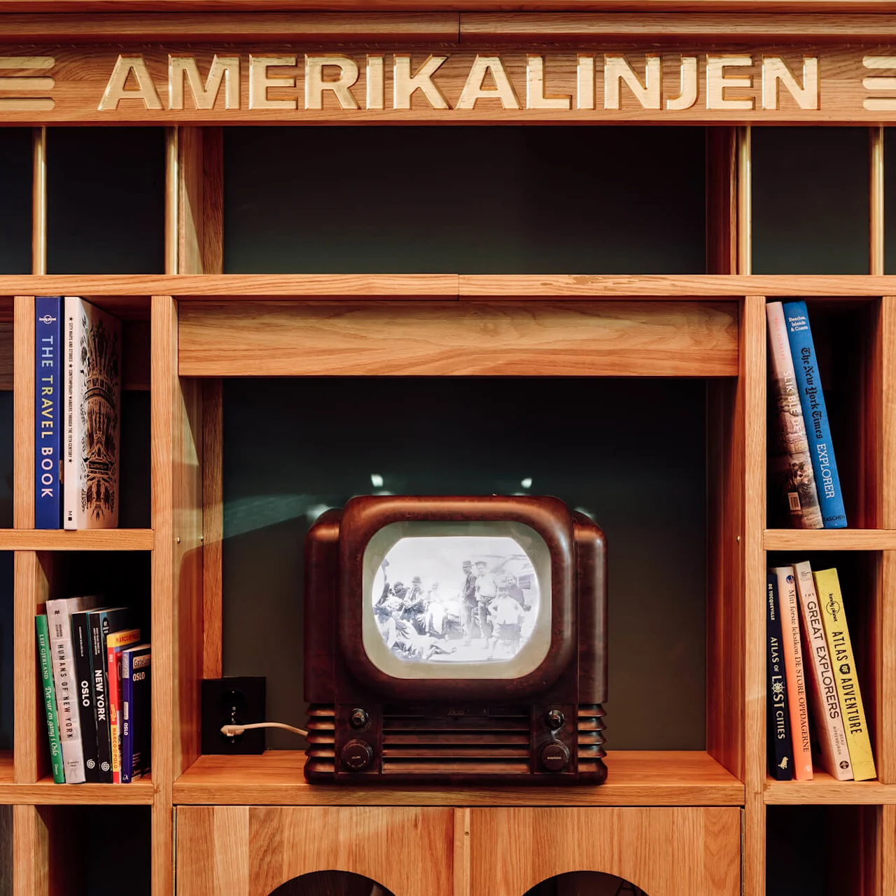 A vintage TV displaying a black-and-white image sits on a wooden bookshelf filled with books. "AMERIKALINJEN" is written above. Books include "THE TRAVEL BOOK" and "NEW YORK."
