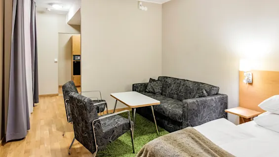 Et lite oppholdsrom med en svartmønstret sofa og to stoler rundt et hvitt kaffebord. Rommet har lyse vegger og tregulv, med myk belysning.