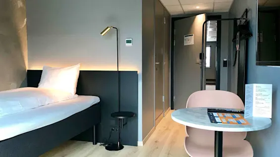 Et pænt arrangeret hotelværelse har en enkeltseng, en stålampe og et lille bord med en stol. En menu på bordet tilbyder forskellige tilbehør.