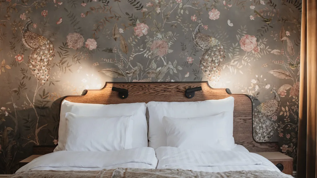 Home Hotel Baltzar - Deluxe Double. A cozy bed with a wooden headboard and elegant peacock and floral wallpaper.