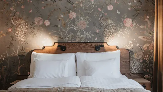 Home Hotel Baltzar - Deluxe Double. A cozy bed with a wooden headboard and elegant peacock and floral wallpaper.