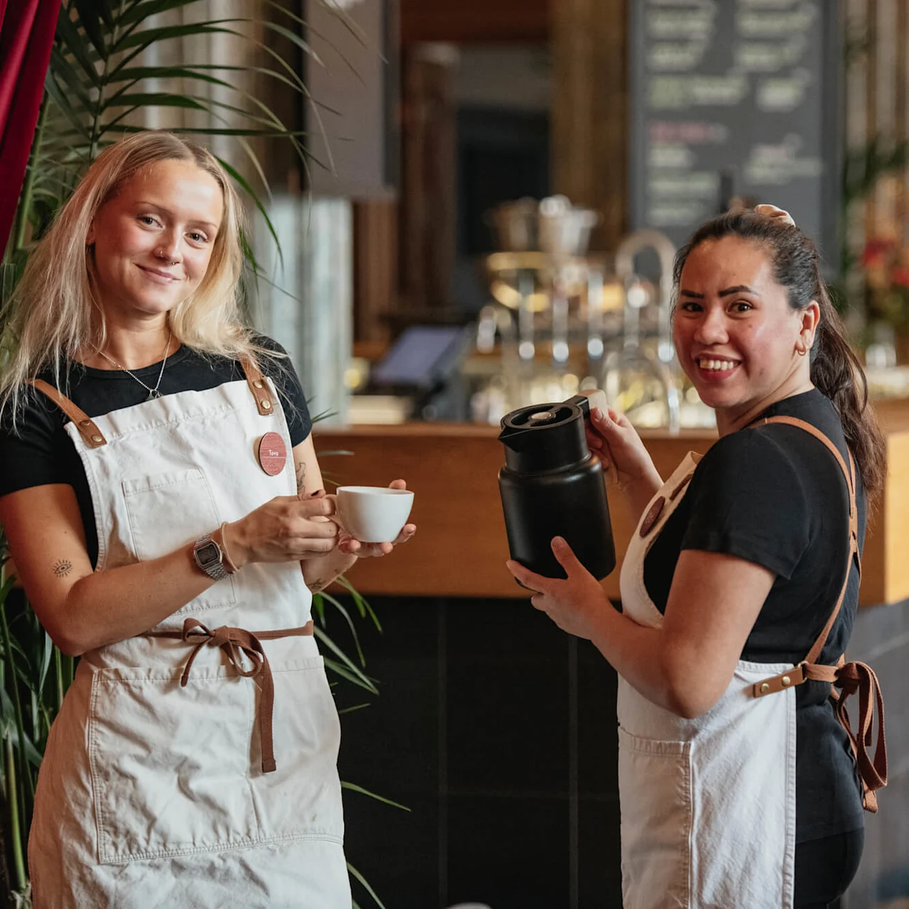 To smilende baristaer serverer kaffe innendørs, en med en kopp og den andre med en termos.