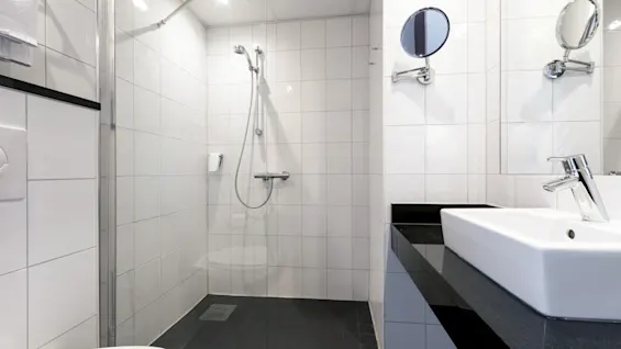 Moderni kylpyhuone, jossa lasinen suihku, kromiset varusteet ja valkoinen pesuallas mustalla tasolla. Laatoitetut seinät ja lattia korostavat puhdasta, minimalistista suunnittelua.