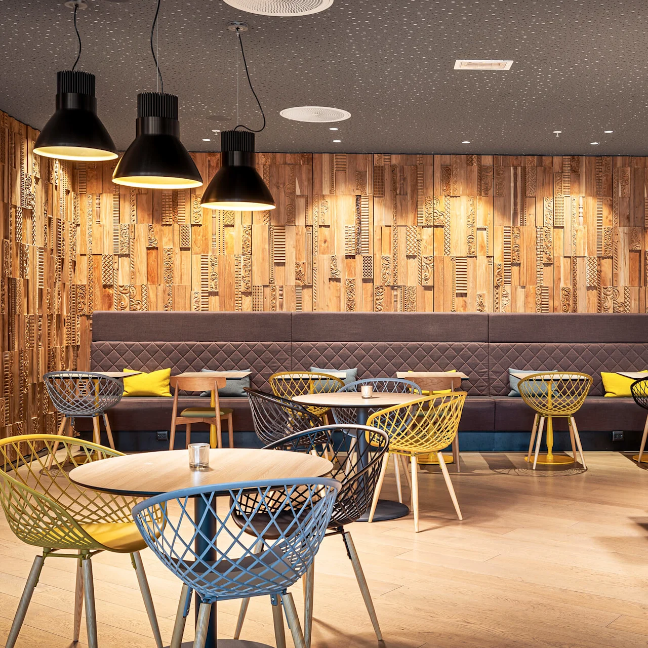 A café setting with modern chairs surrounding round tables, under large black pendant lights. The wall features intricate wooden paneling, and the floor is light wood.