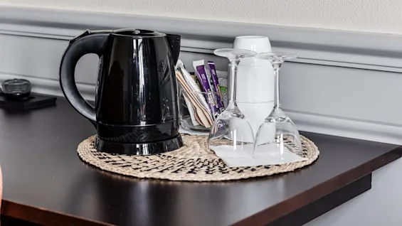 A black electric kettle, packets of sugar, and upside-down glasses on a woven mat, on a dark wood surface.