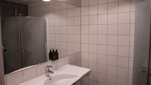 Handfat och bänkskiva med toalettartiklar och glas i ett kaklat badrum med dusch i hörnet. Överliggande lampor lyser upp scenen.