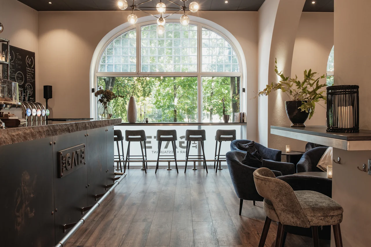 Interiør af en bar og lounge med et stort buet vindu, der viser grønne træer. Barstole og lænestole. FGATE RESTAURANT.