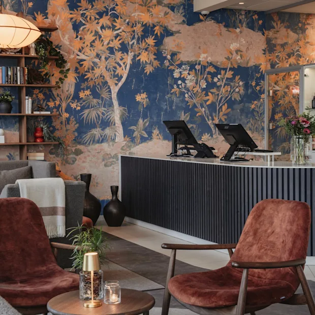 En hotellounge med servicedisk, bar og et hyggeligt siddeområde. Et levende vægmaleri af træer og løvværk pryder væggen.