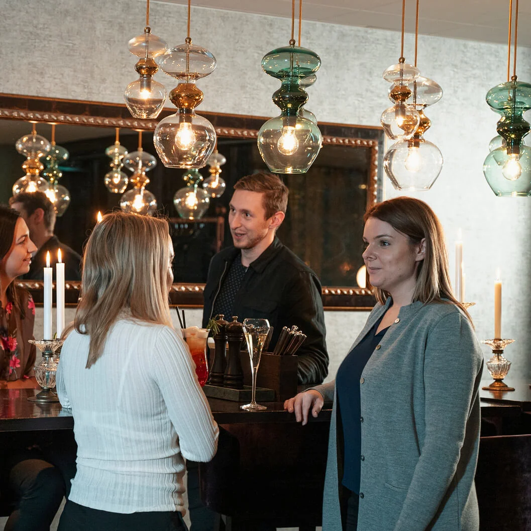 Personer, der taler omkring et bardiskbord, med farverige cocktails. De befinder sig i et varmt oplyst rum med hængende lamper og et stort spejl, der reflekterer scenen.