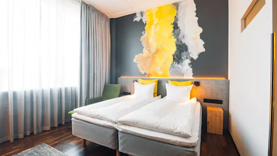 Twin beds with white linens sit in a modern room featuring a vibrant yellow and grey mural on the wall, next to a large window with grey curtains.