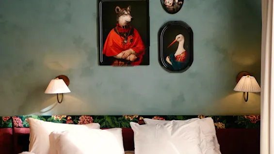 A neatly made bed with white sheets and pillows sits beneath three animal portraits in ornate frames. Soft wall lamps are on either side, in a room with teal walls and floral accents.