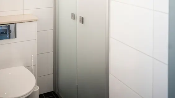 Ett badrum har vita kakelväggar och en glasduschdörr bredvid en toalett. En metallisk spolknapp är på väggen. Utrymmet är rent och modernt.