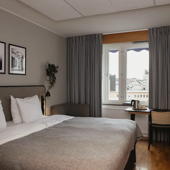 En fint bäddad säng med grå sängkläder står i ett hotellrum. Till höger står ett skrivbord med en vattenkokare och lampa, med ett fönster mot stadens tak.