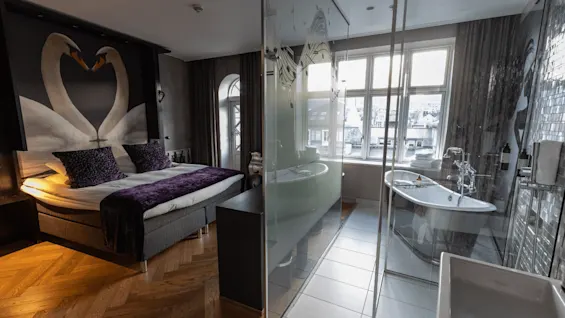 Et koselig soverom med dobbeltseng, lilla puter og en svanemaleri. Ved siden av er et moderne bad med glassvegger og frittstående badekar, badet i naturlig lys fra store vinduer.
