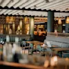 A modern restaurant features green velvet seating and wooden tables. Warm pendant lights create an inviting atmosphere. A laptop and cup are on one table.
