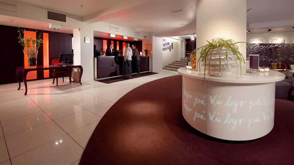 Et hotellobby med gæster, der tjekker ind, med receptionsdisk, siddeområde og dekorativ belysning.