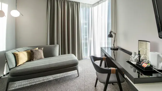 Et moderne hotellrom har en grå chaiselong med puter, et skrivebord med lampe og serveringsbrett, og gardiner fra gulv til tak som filtrerer sollys.