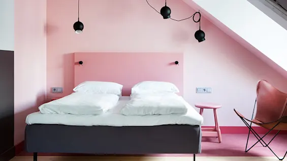 En seng med hvitt sengetøy står i et rosa malt rom, med svarte hengelamper over. En rosa krakk og brun stol fullfører den minimalistiske innredningen.