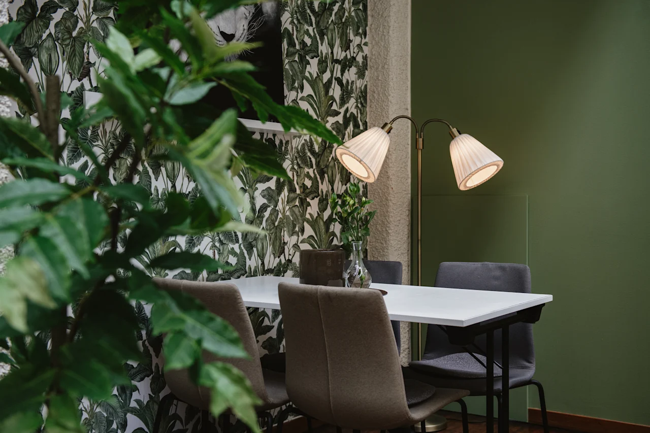Vitt bord med beige stolar, upplyst av en lampa med två skärmar, i ett rum med gröna väggar och bladig tapet; en växt syns i förgrunden.