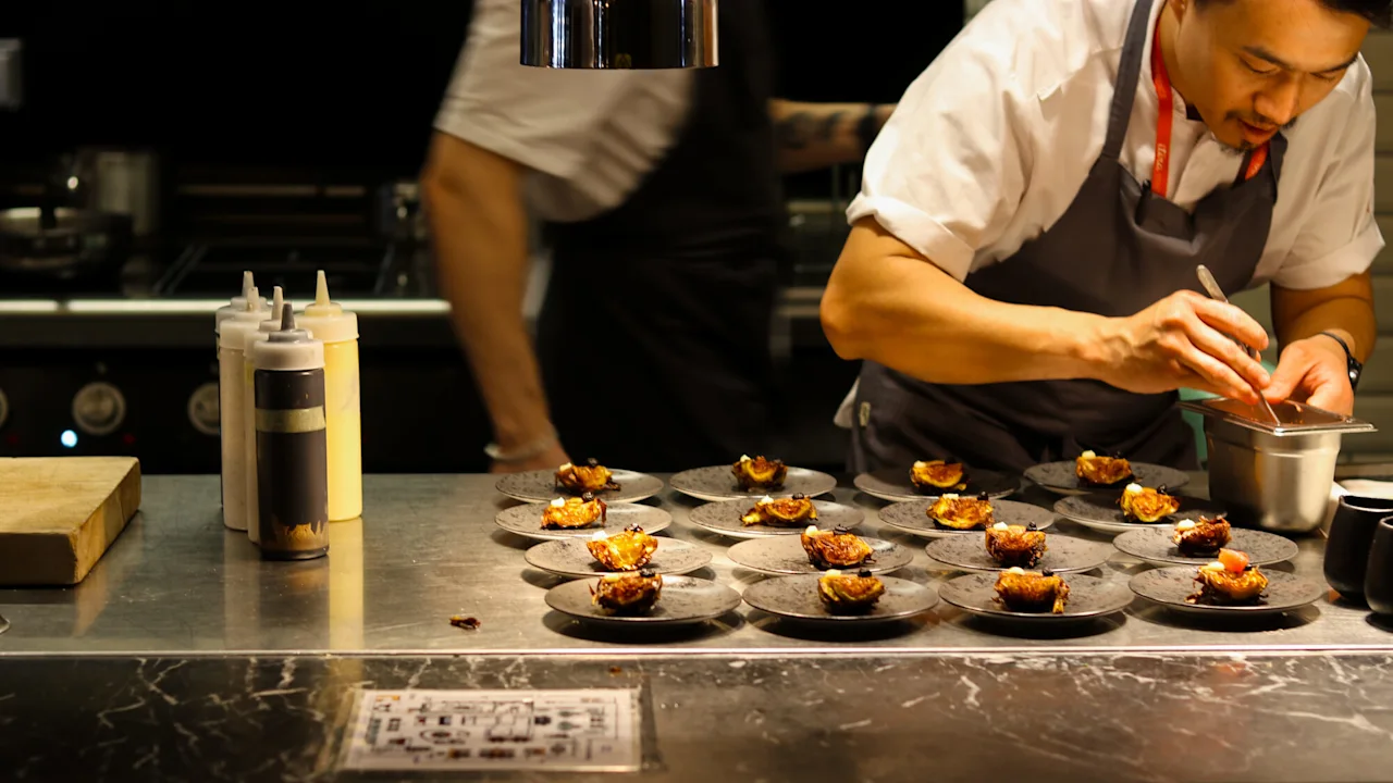 Kokk pynter anrett mat på tallerkener på et kjøkken av rustfritt stål og tilbereder flere små porsjoner. Flasker og skjærebrett er i nærheten, som skaper en travel, profesjonell kulinarisk atmosfære.