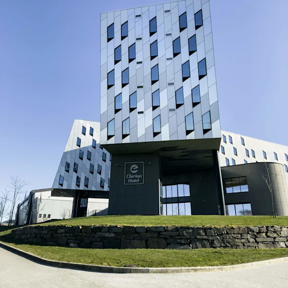 En moderne bygning med geometriske mønstre og mange vinduer står mot en klar himmel. Skilt med teksten "Clarion Hotel". Omgivelsen inkluderer gress, steinmur og trær.