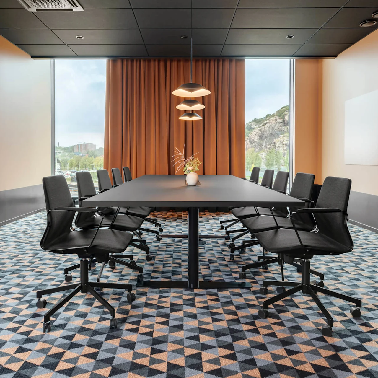 Ett långt svart konferensbord med svängbara stolar runt det, placerat i ett modernt mötesrum med mönstrad matta, orange gardiner, stora fönster och pendellampor ovanför.