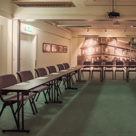 Rectangular tables arranged in U-shape; conference room setting with chairs, papers, and a projector. Interior with art and warm lighting.