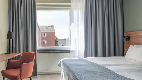 En prydligt bäddad säng med grå filt står bredvid ett fönster, delvis täckt av mörka gardiner, med utsikt över närliggande hustak. En brun stol och lampa vid ett skrivbord i trä.