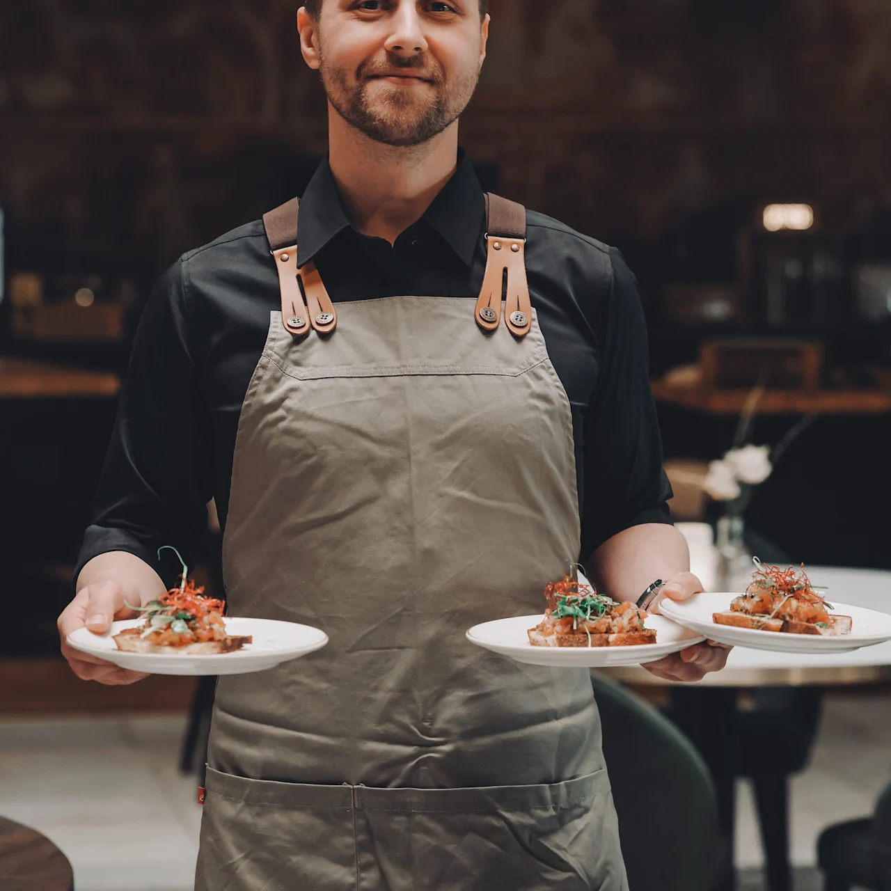 A waiter carries three plates of appetizing bruschetta, wearing a gray apron in an upscale restaurant setting with dark, elegant decor.