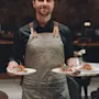 A waiter carries three plates of appetizing bruschetta, wearing a gray apron in an upscale restaurant setting with dark, elegant decor.