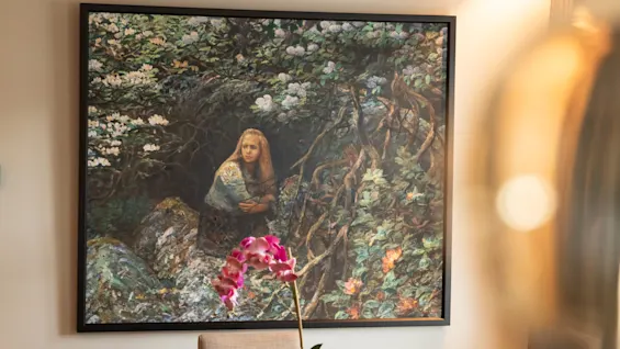 A round table with beige chairs surrounds a potted orchid. A painting, depicting a girl in a forest scene, is mounted on the wall behind.