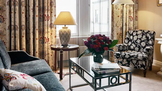 En stue med en blomsterlænestol og sofa, et bord med lamper, et sofabord med blomster og magasiner, alt sat mod blomstrede gardiner og et vindue.