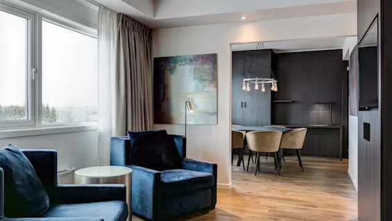 Moderni hotellisviitti, jossa on siniset samettinojatuolit, puulattiat ja taustalla näkyvä ruokailutila.