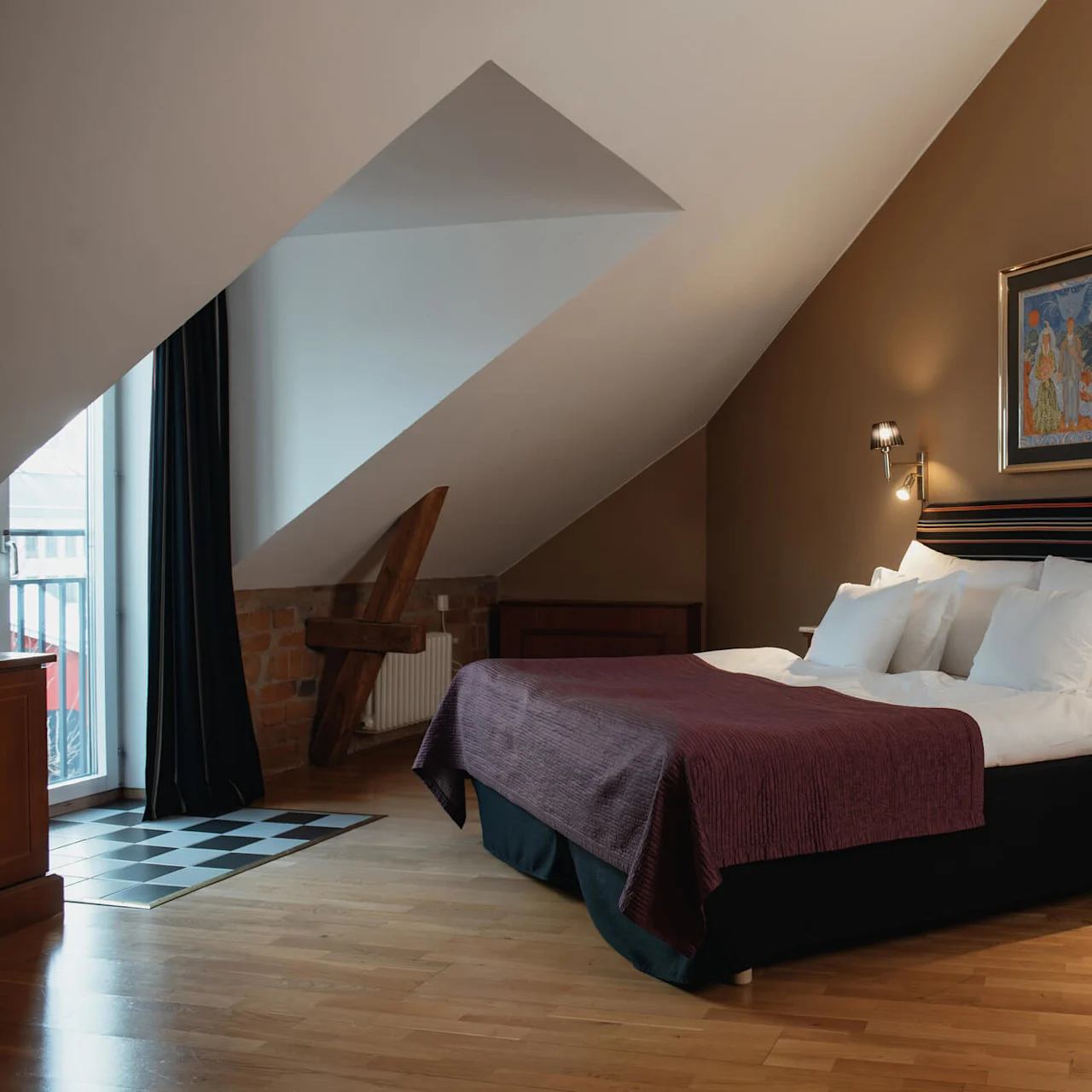 A large bed with multiple pillows and a purple bedspread sits under a sloped ceiling. Two wall lamps illuminate a painting, while wooden furniture and brickwork create a cozy atmosphere.