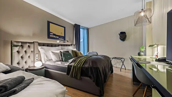 Bed with pillows and blanket situated in a modern bedroom; walls are adorned with artwork and a decorative hat. A desk with a lamp and plant is nearby.