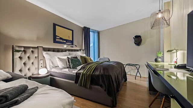 Seng med puder og tæppe placeret i et moderne soveværelse; væggene er dekoreret med kunst og en dekorativ hat. Et skrivebord med en lampe og plante er i nærheden.