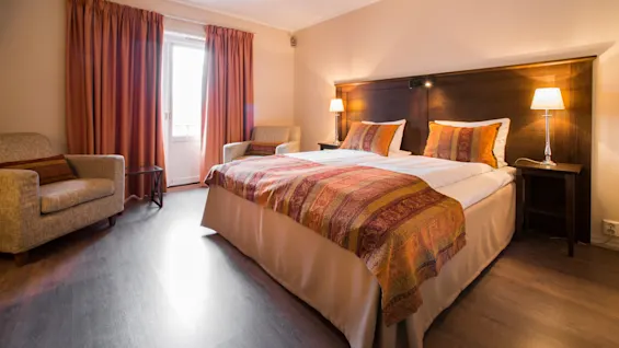 En snyggt bäddad säng med färgglad bäddning i ett välbelyst hotellrum med fåtöljer och trägolv.