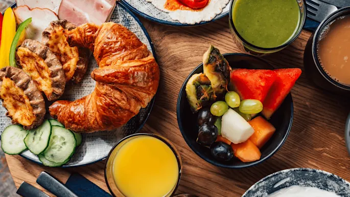 Croissant, skinke og osteboller ligger på et bord med skivede grønnsaker, fruktsalat, appelsinjuice, grønn smoothie og kaffe rundt.