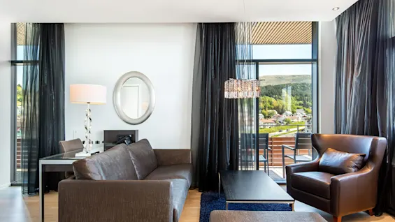 Et moderne oppholdsrom har en brun sofa vendt mot et glassbord. En skinnstol og gulvlampe står ved siden av mørke gardiner, med naturskjønn fjellutsikt utenfor store vinduer.