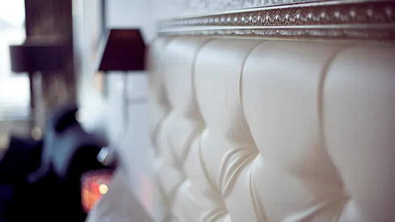 A white tufted headboard with intricate detailing against a blurry room backdrop.