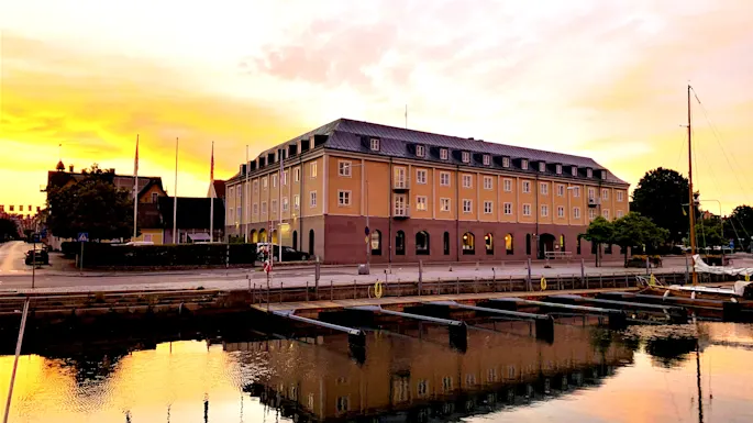 En hotellbyggnad vid en hamn som speglar sig i vattnet under en gyllene solnedgång.