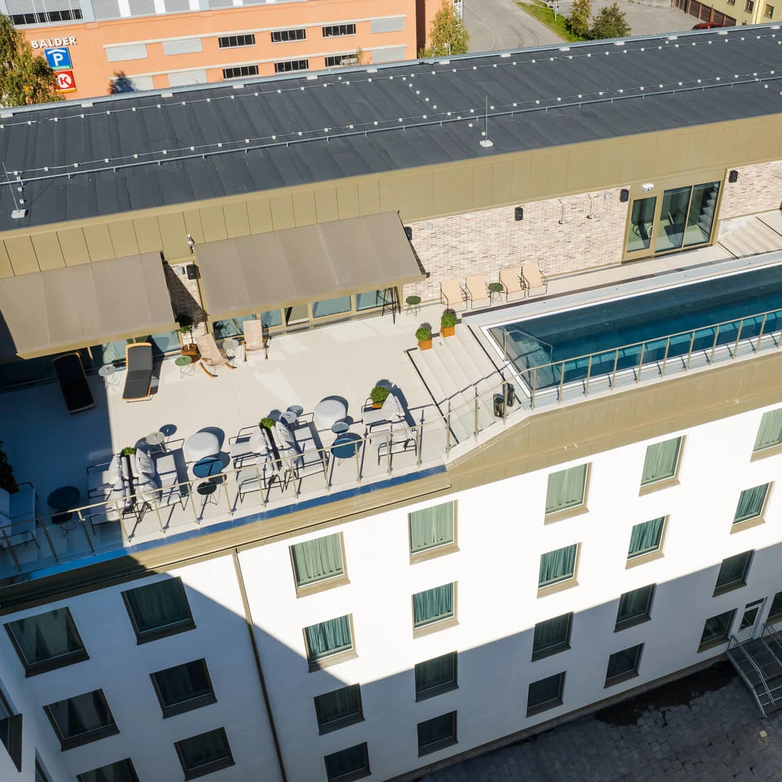 En tagterrasse har en rektangulær pool og siddepladser, omgivet af en moderne bygning. I nærheden er der et skilt med “BALDER P” og anden uklar skiltning på en tilstødende væg.