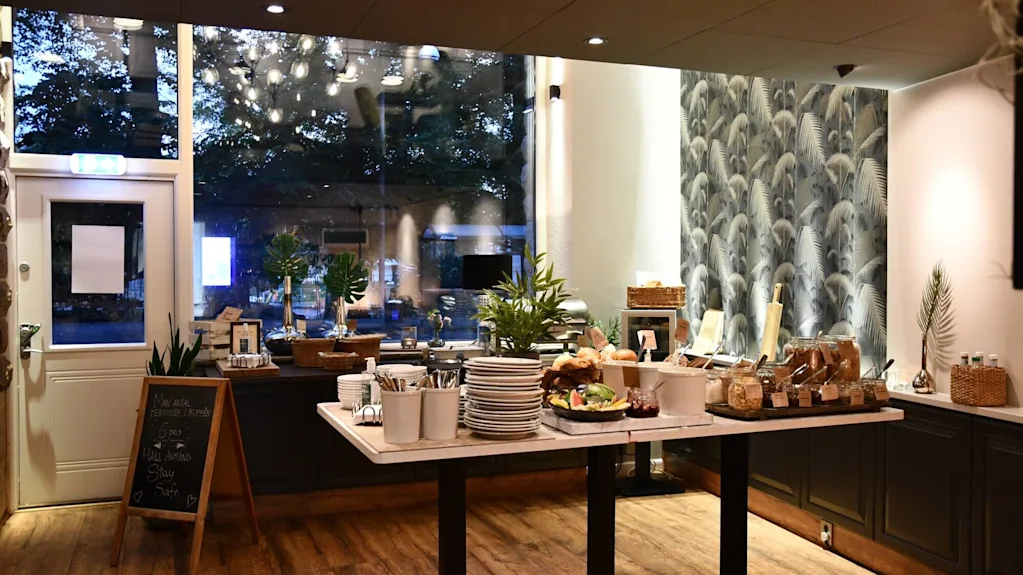 Et buffébord med mat og tallerkener inne i et lysfylt rom med store vinduer og tapet med bladmønster.
