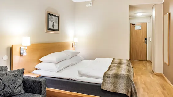 En pent oppredd seng er sentrert i et hotellrom, flankert av to vegglamper. Et abstrakt maleri henger over trehodegjerdet, og en lenestol står like ved.