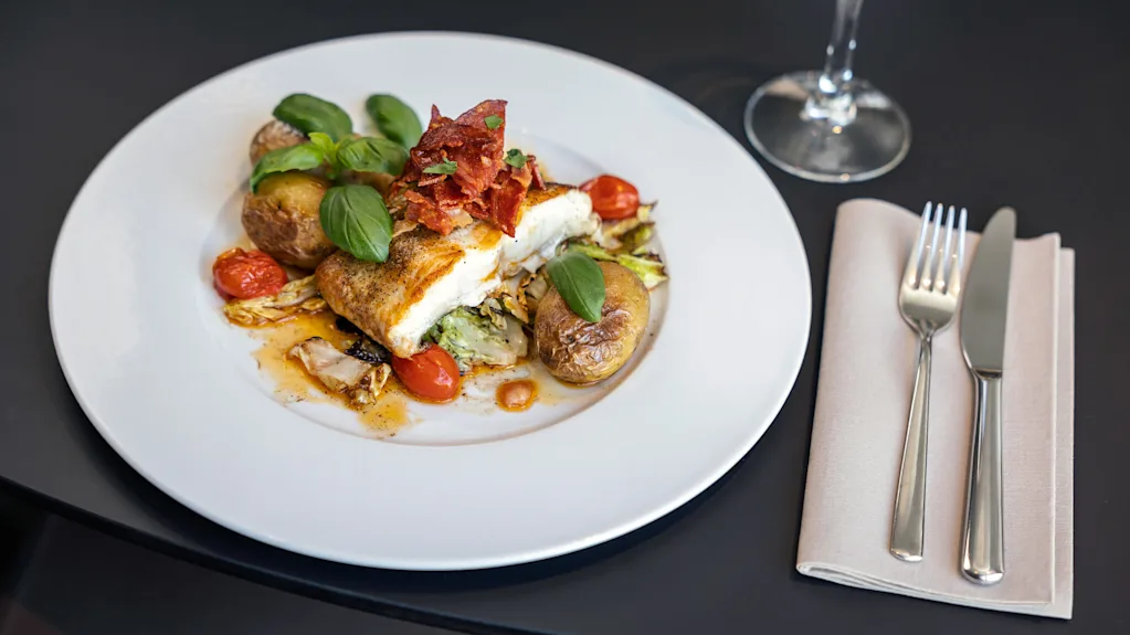 A plated dish of fish garnished with leafy greens and cherry tomatoes, accompanied by roast potatoes, sits on a table beside silverware and an empty wine glass.