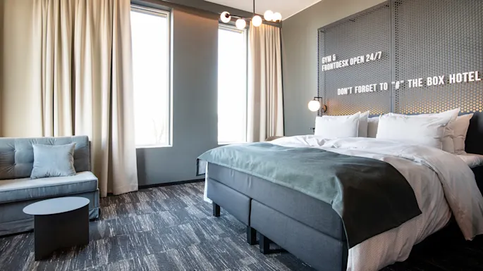 En pent oppredd seng med hvite laken står i et moderne hotellrom. Hvite gardiner og vegger skaper en lys atmosfære. Teksten lyder: "GYM & FRONTDESK OPEN 24/7. DON'T FORGET TO '#' THE BOX HOTEL."